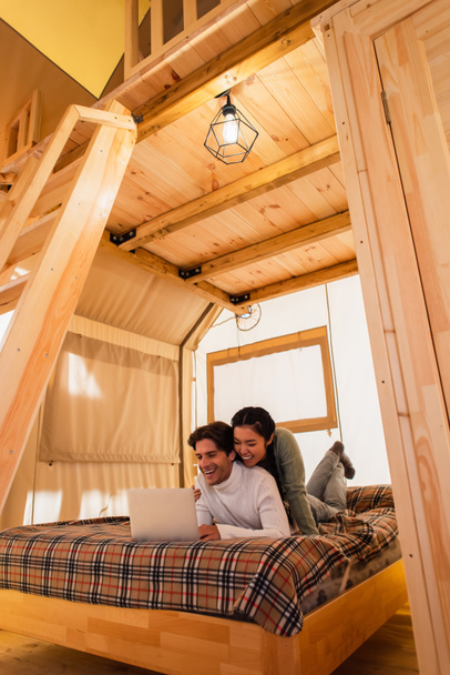 Smiling interracial couple using laptop on bed in wooden glamping house  - Фото, изображение