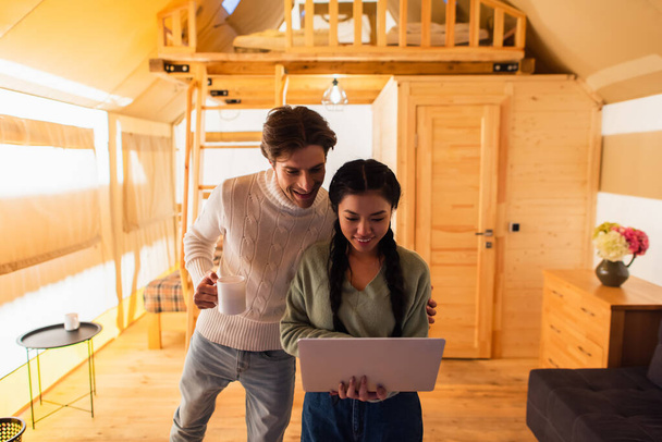 Interracial couple with cup using laptop in glamping house during weekend  - Foto, Imagen