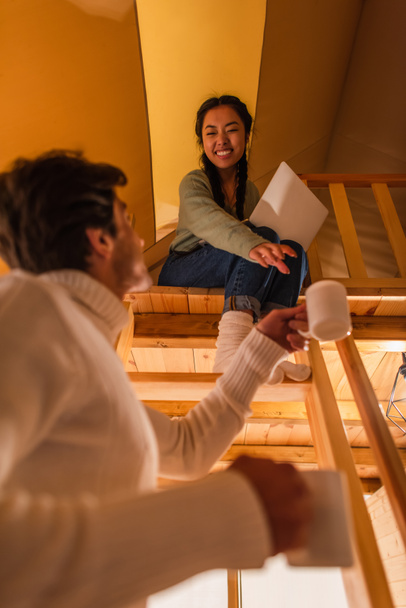 Low angle view of man holding cups near asian girlfriend with laptop in glamping house  - Photo, Image