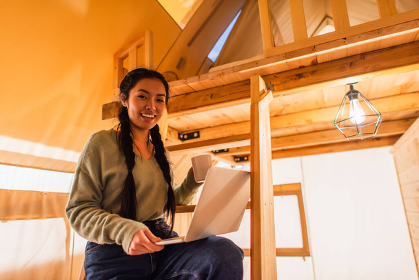 Asian woman with cup and laptop looking at camera in glamping house  - Foto, immagini