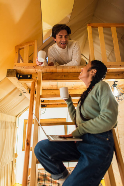 Low angle view of smiling interracial couple with cups and laptop looking at each other in glamping house  - Fotoğraf, Görsel