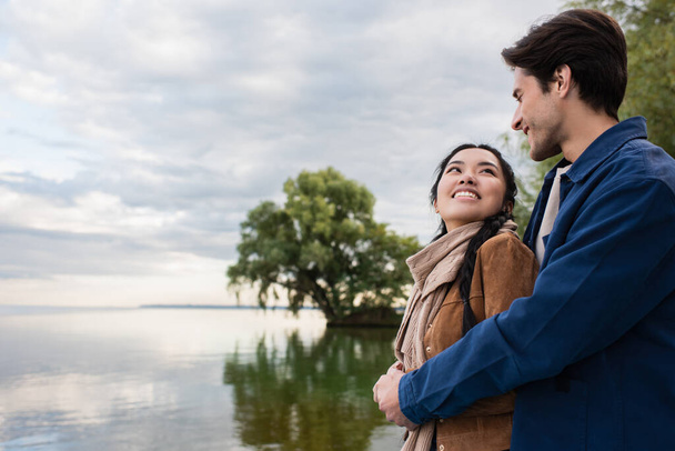 Smiling man hugging asian girlfriend near lake during vacation  - Foto, imagen
