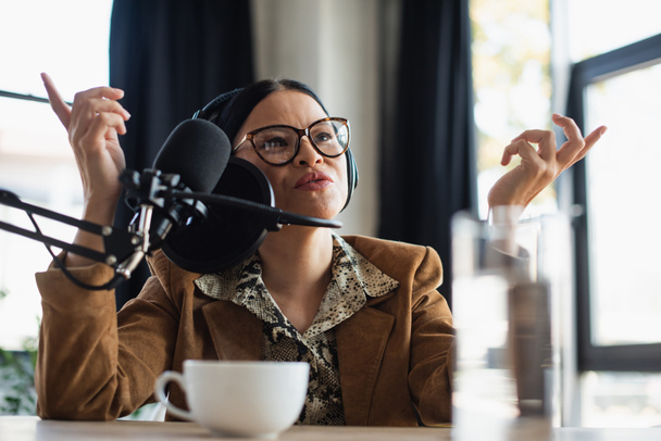 asiatique radio hôte dans lunettes et écouteurs gestuelle près tasse de café sur le bureau - Photo, image