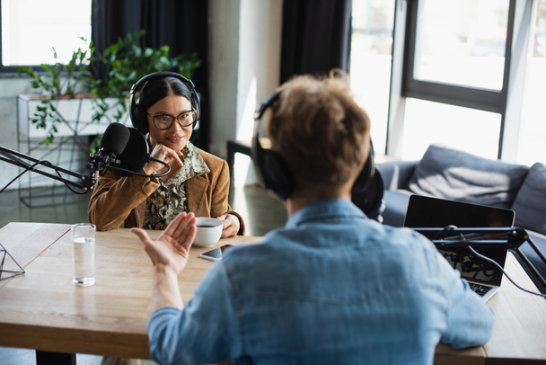 feliz asiático rádio host em óculos e fones de ouvido olhando para confuso colega durante podcast - Foto, Imagem