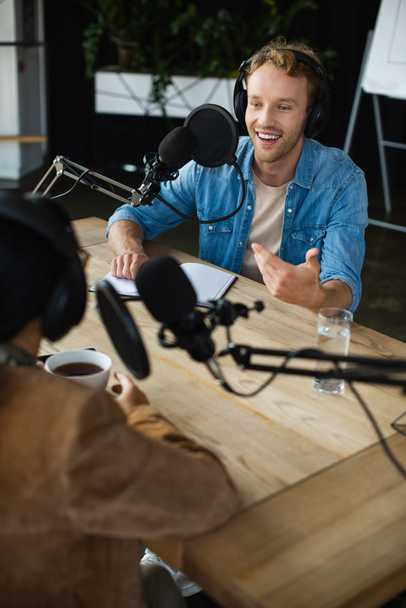 χαρούμενο ραδιοφωνικό ξενιστή σε ακουστικά κοιτάζοντας θολή συνάδελφο και gesturing κατά τη διάρκεια podcast - Φωτογραφία, εικόνα