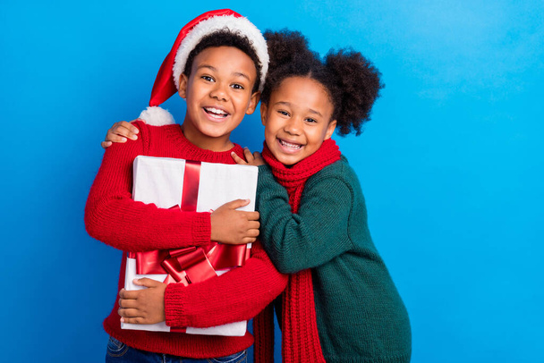 Foto di divertente dolce pelle scura bambini vestiti maglioni copricapo sciarpa tenendo regalo sorridente abbraccio isolato colore blu sfondo - Foto, immagini