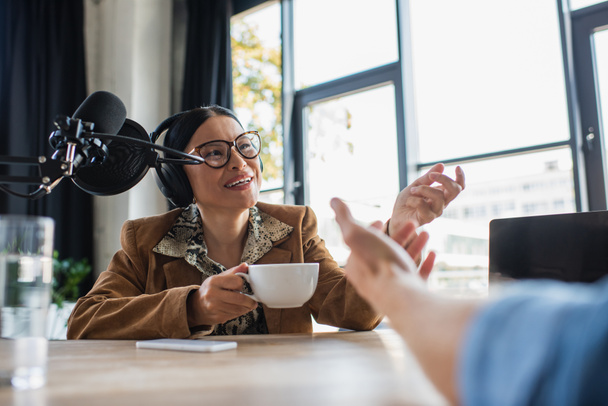 alegre asiático rádio host no óculos e fones de ouvido falando com colega e segurando copo durante podcast - Foto, Imagem