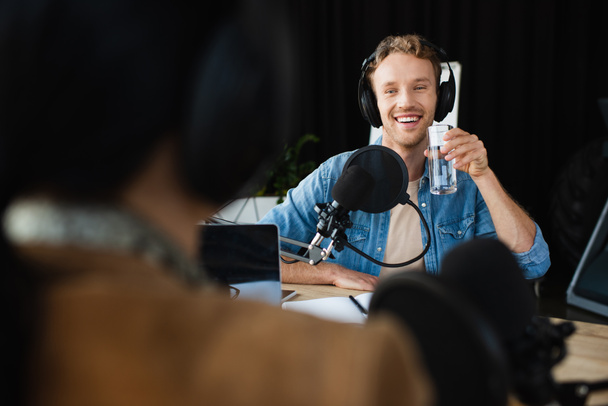 happy radio host in headphones looking at blurred colleague and holding glass of water during podcast - Фото, изображение