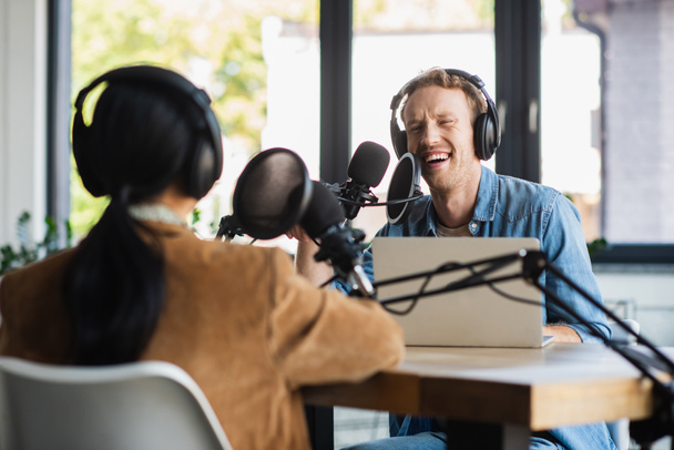 χαμογελαστός ραδιοφωνικός ξενιστής σε ακουστικά μιλώντας στο μικρόφωνο και κοιτάζοντας θολή συνάδελφο κατά τη διάρκεια podcast - Φωτογραφία, εικόνα