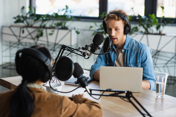 ραδιοφωνικός ξενιστής σε ακουστικά μιλώντας στο μικρόφωνο κοντά στο laptop και θολή συνάδελφο κατά τη διάρκεια podcast - Φωτογραφία, εικόνα