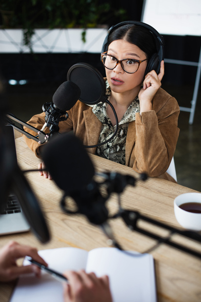 surpresa asiático rádio host em óculos e fones de ouvido falando e olhando para confuso colega  - Foto, Imagem