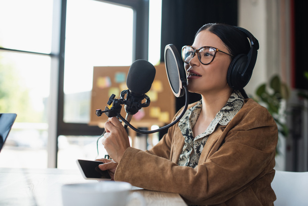 Asijské rádio hostitel v brýlích a sluchátka drží smartphone při výrobě podcast  - Fotografie, Obrázek
