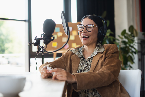 positif asiatique radio hôte dans lunettes et écouteurs tenant smartphone tout en riant pendant podcast  - Photo, image