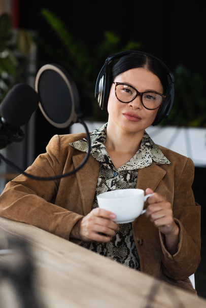 Asijské rádio hostitel v brýlích a sluchátka drží šálek kávy a dívá se do kamery - Fotografie, Obrázek