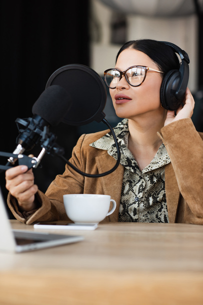 Ασιάτης ραδιοφωνικός παρουσιαστής σε γυαλιά και ακουστικά που μιλάει κοντά σε φλιτζάνι καφέ κατά τη διάρκεια του podcast  - Φωτογραφία, εικόνα
