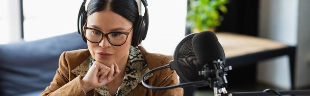 pensieroso host radio asiatica in occhiali e cuffie vicino al microfono con filtro pop durante il podcast, banner - Foto, immagini