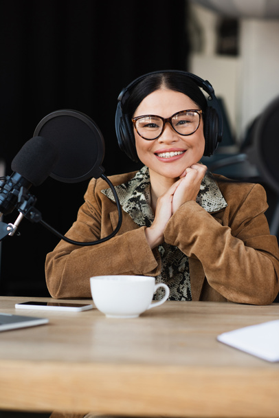 heureux asiatique radio hôte dans lunettes et écouteurs sourire près de tasse et smartphone sur le bureau - Photo, image