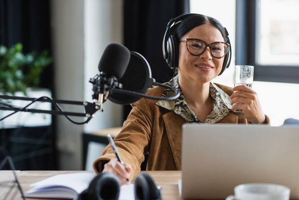 pozitivní asijské rádio hostitel ve sluchátkách drží sklenici vody a pera v blízkosti notebooku ve studiu - Fotografie, Obrázek