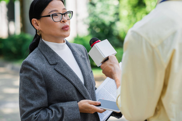 репортер держит микрофон рядом с азиатской бизнесвумен в очках со смартфоном  - Фото, изображение