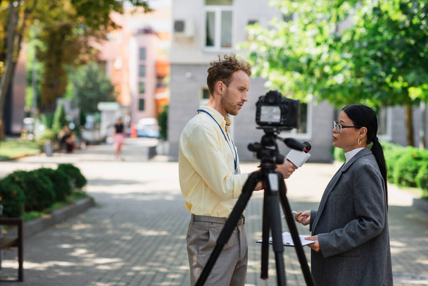 vista lateral de newscaster segurando microfone perto asiático empresária em óculos na rua  - Foto, Imagem