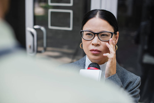 θολή δημοσιογράφος με μικρόφωνο κοντά σε σκεπτική ασιατική επιχειρηματίας προσαρμογή γυαλιά  - Φωτογραφία, εικόνα