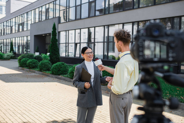 記者がマイクを持ってビルの近くに立っている眼鏡で陽気なアジア人のビジネスマンのインタビューを受け  - 写真・画像
