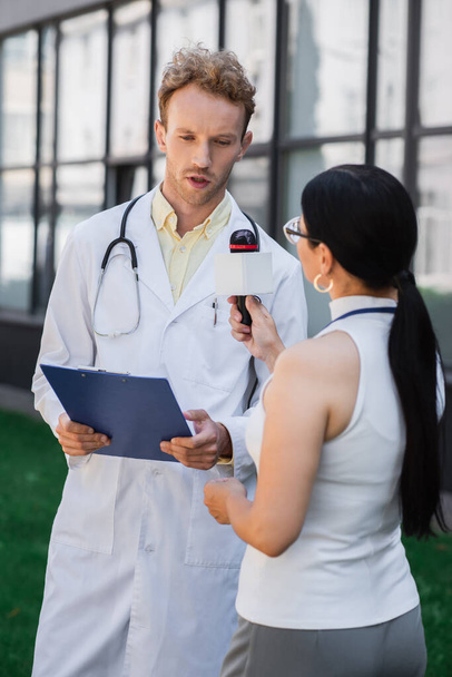 brunette asiatique journaliste tenant microphone près médecin en manteau blanc - Photo, image