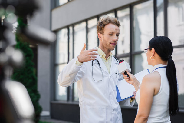 γιατρός με λευκό παλτό κρατώντας πρόχειρο και gesturing, ενώ δίνει συνέντευξη σε ασιάτης δημοσιογράφος με μικρόφωνο  - Φωτογραφία, εικόνα