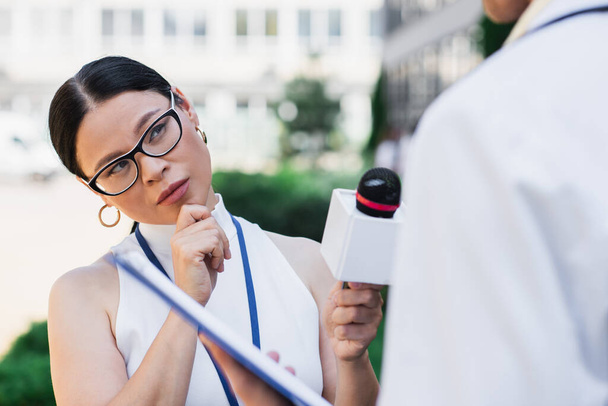 brunetka asijský novinář držení mikrofon a při pohledu na rozmazané lékaře v bílém plášti  - Fotografie, Obrázek
