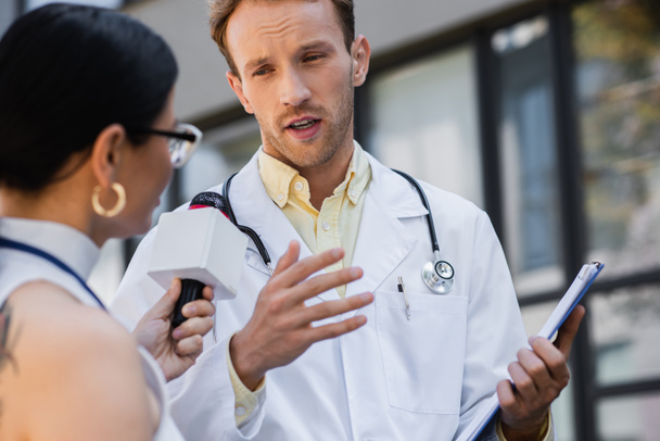 doctor in white coat holding clipboard and gesturing while giving interview to blurred and tattooed asian journalist with microphone  - Foto, Imagen