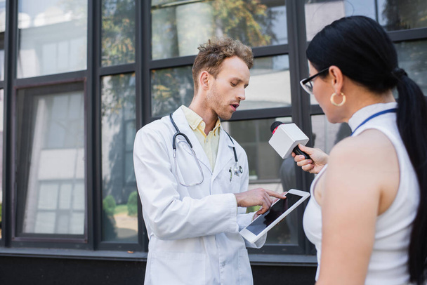врач в белом халате указывает на цифровой планшет, давая интервью размытому азиатскому журналисту с микрофоном  - Фото, изображение