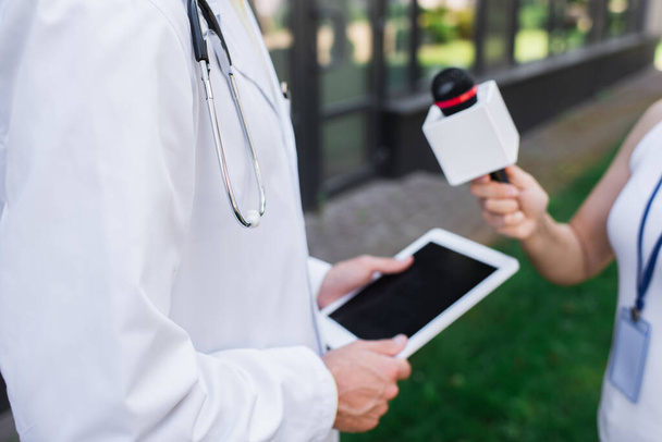 przycięty widok lekarza w białym płaszczu trzymającego cyfrową tabletkę podczas rozmowy z dziennikarzem z mikrofonem  - Zdjęcie, obraz