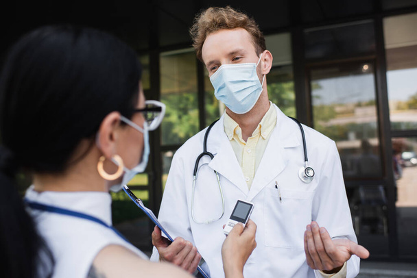 borrosa asiático reportero celebración voz grabadora cerca médico en médico máscara y blanco abrigo  - Foto, Imagen