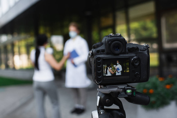 cámara digital con reportero asiático sosteniendo micrófono cerca del médico en máscara médica durante la entrevista en la pantalla - Foto, Imagen