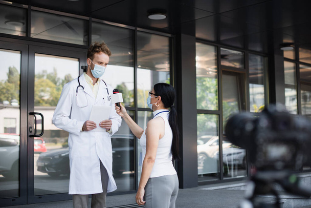 reporter asiatique dans masque médical tenant microphone près du médecin en manteau blanc avec tablette numérique debout près de la clinique - Photo, image