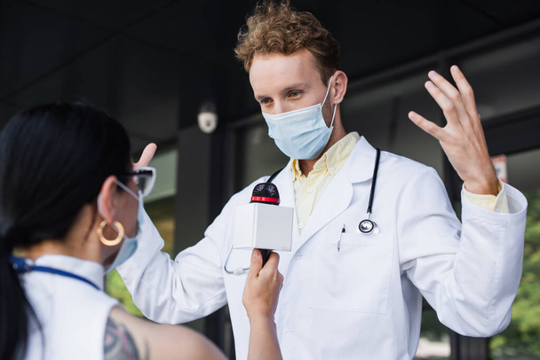 doctor in medical mask gesturing during interview with blurred asian reporter - Φωτογραφία, εικόνα