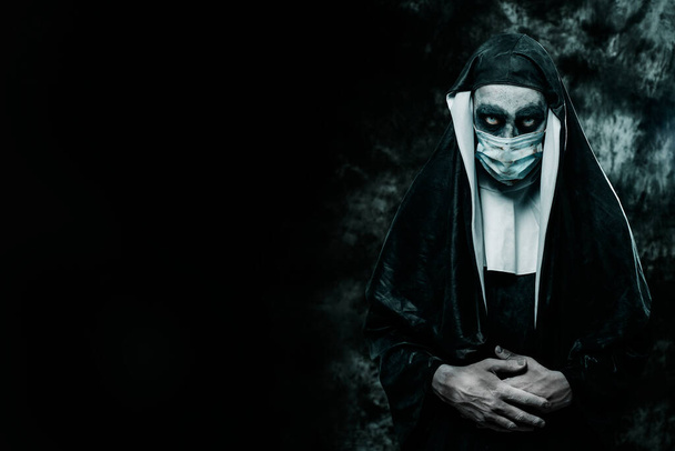 uma freira assustadora do mal, vestindo um hábito típico preto e branco e uma máscara facial descartável, em um fundo preto com algum espaço em branco à esquerda - Foto, Imagem