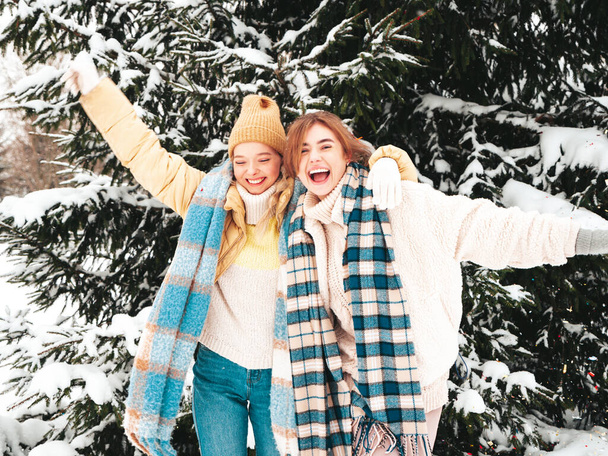 Two young beautiful smiling hipster female in trendy warm clothes and scarfs.Carefree women posing in the street in park. Positive pure models having fun in snow. Enjoying winter moments. Xmas concept - Foto, Imagen