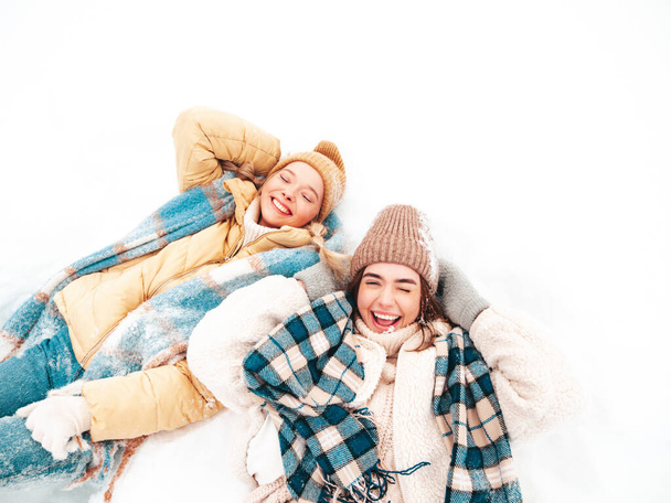 Two young beautiful smiling hipster female in trendy warm clothes and scarfs.Carefree women posing in the street in park. Positive pure models lying in snow. Enjoying winter moments. Xmas concept - Photo, image