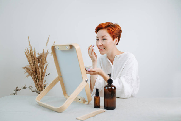Lively short-haired woman looking at the mirror with a wooden frame, applying the concealer. Using eco-friendly products and things. - Valokuva, kuva