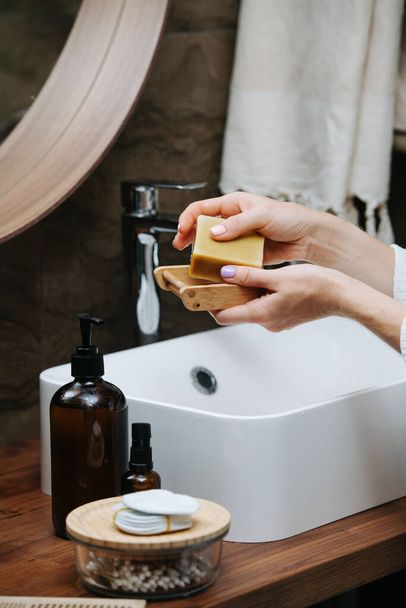 Wooden soap dish in hands of a mature woman with an eco soap block. She's standing in front of the sink in a bathroom. - Photo, image