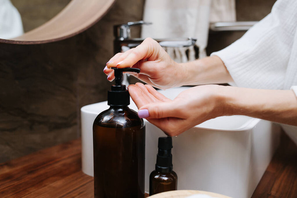 Close up image of hands of a mature woman pumping an eco soap from a bottle. She's standing in front of the sink in a bathroom. - Foto, afbeelding