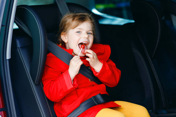 Μικρό κοριτσάκι κάθεται στο κάθισμα του αυτοκινήτου, κρατώντας γλυκό γλειφιτζούρι ενάντια στη ναυτία και την ασθένεια και κοιτάζοντας έξω από το παράθυρο για τη φύση και την κυκλοφορία. Χαριτωμένο παιδί που ταξιδεύει με αυτοκίνητο. Ασφάλεια των παιδιών στο δρόμο. - Φωτογραφία, εικόνα