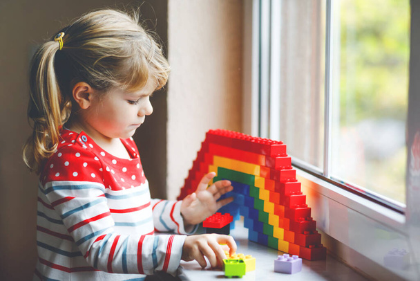 Cute little toddler girl by window create rainbow with colorful plastic blocks during pandemic coronavirus quarantine. Children made and paint rainbows around the world as sign. - Foto, imagen