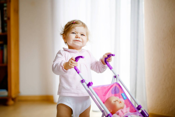 Söpö suloinen vauva tyttö tekee ensimmäiset askeleet nukkevaunun kanssa. Kaunis lapsi työntää rattaita lelulla kotona. Onnellinen tytär oppii kävelemään ja seisomaan - Valokuva, kuva