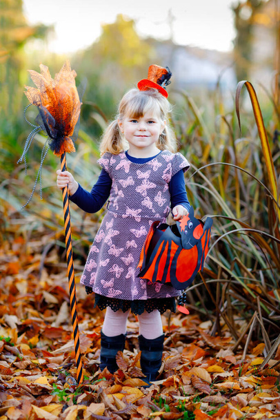 Aranyos kislány boszorkánynak öltözve ünnepli a Halloweent. Boldog gyerek kint, narancssárga vicces kalapban, boszorkányseprűvel és denevérlámpás táskával. Gyönyörű családi fesztivál szezon októberben. - Fotó, kép