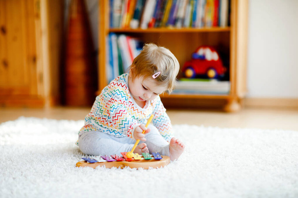 家庭や保育園で教育的な木製の音楽のおもちゃで遊んでいる小さな赤ちゃんの女の子。カラフルな木製のキシロフォンと幼児の子供。コロナウイルス感染症の隔離中の孤独な子供. - 写真・画像