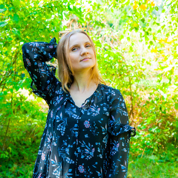 Jeune femme blonde dans une robe noire avec un imprimé floral dans le parc d'automne, journée ensoleillée. Mode de rue, style de rue. Image de tenue d'automne - Photo, image
