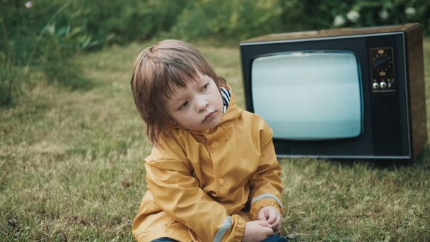 悲しい子供レインコートに身を包んだ草の上に座っている。彼の後ろにレトロテレビを働いて - 写真・画像