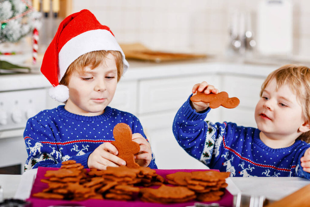 Zwei kleine Vorschulkinder backen Lebkuchen. Glückliche Geschwister, Kinder in Weihnachtspullovern. Weihnachtlich dekorierte Küche. Brüder, die kämpfen und Chaos stiften. Familienaktivitäten zu Weihnachten - Foto, Bild